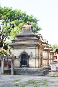 A stone temple structure adorned with intricate carvings and a bell hanging from a wooden frame. The building features ornate, arched windows with lattice designs and is surrounded by lush green trees. The area around the temple is paved with stone slabs, some of which have grass growing between them.