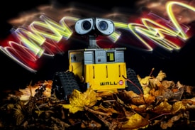 A small robot with a yellow body and large eyes stands among dried leaves. Behind it, colorful light trails swirl against a dark background, creating a vibrant contrast.