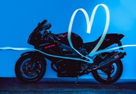 A black motorcycle is depicted on a bright blue background with neon pink accents. Light trails form a heart shape above and around the motorcycle, adding a dynamic and artistic effect.