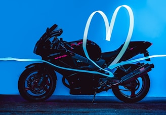 A black motorcycle is depicted on a bright blue background with neon pink accents. Light trails form a heart shape above and around the motorcycle, adding a dynamic and artistic effect.