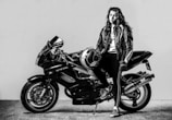 A person wearing a leather jacket stands next to a sleek, powerful motorcycle. The individual holds a helmet and leans confidently against the bike in a black and white setting.