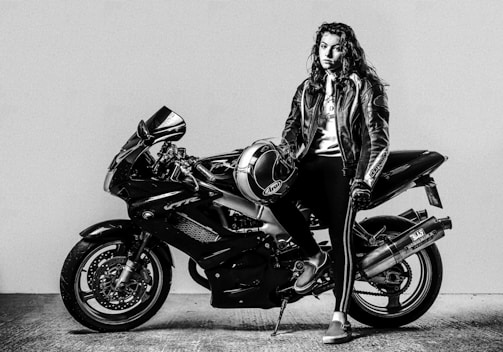 A person wearing a leather jacket stands next to a sleek, powerful motorcycle. The individual holds a helmet and leans confidently against the bike in a black and white setting.