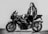 A person wearing a leather jacket stands next to a sleek, powerful motorcycle. The individual holds a helmet and leans confidently against the bike in a black and white setting.