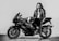A person wearing a leather jacket stands next to a sleek, powerful motorcycle. The individual holds a helmet and leans confidently against the bike in a black and white setting.
