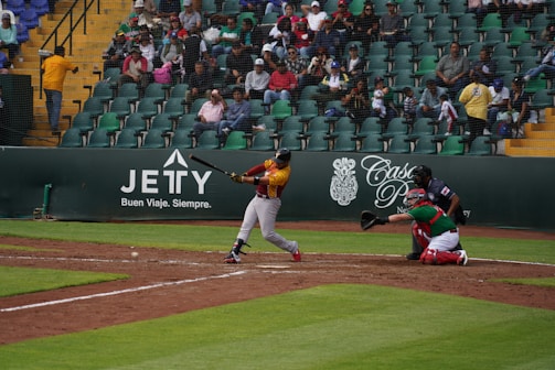 A baseball game in action with a batter swinging at the ball, a catcher in red and green gear crouched behind him, and an umpire observing the play. The field is lined with green and yellow bleachers filled with spectators watching the game.