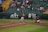 A baseball game in action with a batter swinging at the ball, a catcher in red and green gear crouched behind him, and an umpire observing the play. The field is lined with green and yellow bleachers filled with spectators watching the game.