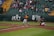 A baseball game in action with a batter swinging at the ball, a catcher in red and green gear crouched behind him, and an umpire observing the play. The field is lined with green and yellow bleachers filled with spectators watching the game.