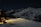 A snow-covered mountain landscape at night, with a cabin on the left side emitting warm, glowing light through the windows. The rugged, snow-laden mountains extend into the distance under a clear, starry sky. A snowmobile is parked near the cabin, and its headlights cast a long beam across the snow.