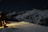A snow-covered mountain landscape at night, with a cabin on the left side emitting warm, glowing light through the windows. The rugged, snow-laden mountains extend into the distance under a clear, starry sky. A snowmobile is parked near the cabin, and its headlights cast a long beam across the snow.