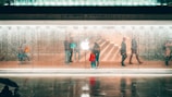 Silhouettes of people walking by a transparent glass facade under a shower of water with a prominent illuminated logo in the center. The interior displays faint reflections and an inviting atmosphere with visible staircases.