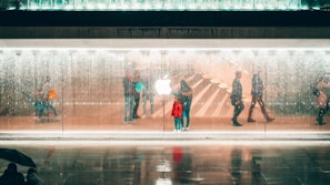 Silhouettes of people walking by a transparent glass facade under a shower of water with a prominent illuminated logo in the center. The interior displays faint reflections and an inviting atmosphere with visible staircases.