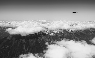 A black and white photograph depicting a landscape where a mountain range is partially obscured by fluffy clouds in the foreground and background. A solitary bird is visible flying in the sky above the mountains, adding a sense of scale and freedom to the scene.