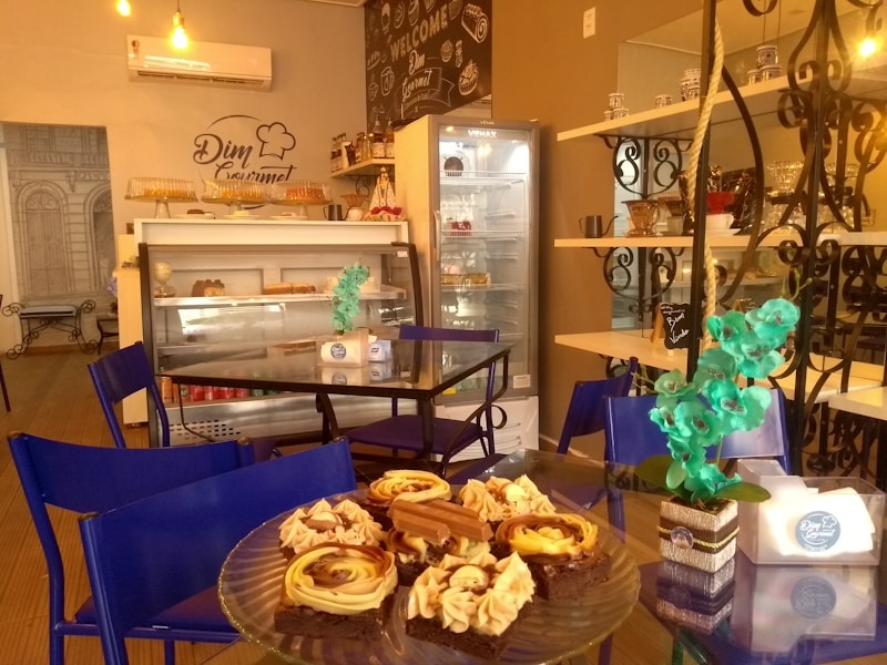 A cozy bakery with a warm, inviting atmosphere. There are tables with blue chairs, a glass display case filled with various pastries and desserts, and a refrigerated display unit in the background. Decorative elements include potted flowers and shelves with jars and decor items. The wall features a chalkboard-style design with the text 'Welcome' and the logo 'Djin Gourmet'.