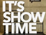 Large white illuminated letters spell out 'IT'S SHOW TIME' against a concrete wall. The letters are mounted on a black frame.