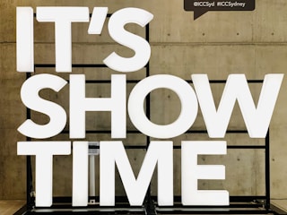 Large white illuminated letters spell out 'IT'S SHOW TIME' against a concrete wall. The letters are mounted on a black frame.