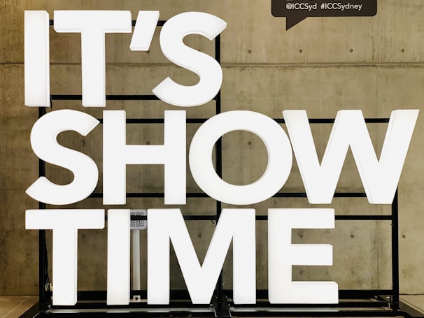 Large white illuminated letters spell out 'IT'S SHOW TIME' against a concrete wall. The letters are mounted on a black frame.