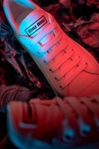 A close-up view of stylish sneakers featuring a gradient lighting effect with hues of red and blue. The sneakers have a label on the tongue and are positioned on a textured surface that resembles crumpled paper.