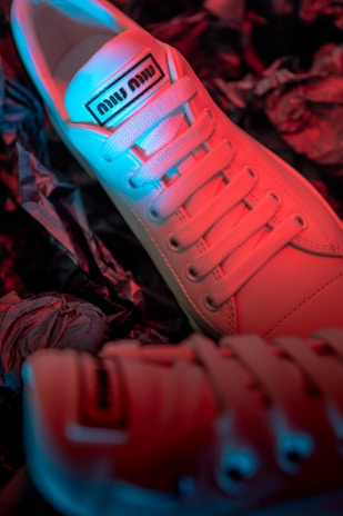 A close-up view of stylish sneakers featuring a gradient lighting effect with hues of red and blue. The sneakers have a label on the tongue and are positioned on a textured surface that resembles crumpled paper.