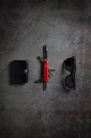 A wallet, a red Swiss Army knife, and a pair of black sunglasses are neatly arranged on a dark, textured surface.