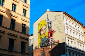 A large mural covers the side of a building, depicting a child wearing a yellow raincoat with animal ears. Next to the child is a whimsical cartoon-like monkey holding a large key. The mural includes the text 'My home might be no palace, but we can share it if you like.' The surrounding walls are part of a cityscape with traditional European-styled architecture under a bright blue sky.