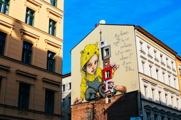 A large mural covers the side of a building, depicting a child wearing a yellow raincoat with animal ears. Next to the child is a whimsical cartoon-like monkey holding a large key. The mural includes the text 'My home might be no palace, but we can share it if you like.' The surrounding walls are part of a cityscape with traditional European-styled architecture under a bright blue sky.