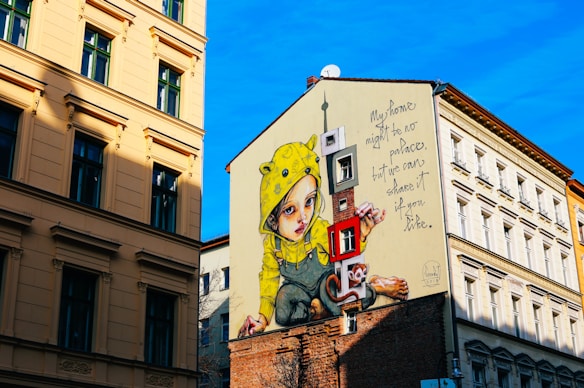 A large mural covers the side of a building, depicting a child wearing a yellow raincoat with animal ears. Next to the child is a whimsical cartoon-like monkey holding a large key. The mural includes the text 'My home might be no palace, but we can share it if you like.' The surrounding walls are part of a cityscape with traditional European-styled architecture under a bright blue sky.