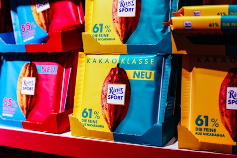 An assortment of colorful Ritter Sport chocolate packages displayed on a shelf. The packages prominently feature percentages indicating the cocoa content and are highlighted with vibrant colors such as blue, red, and yellow. The word 'NEU' is visible, suggesting a new product line.