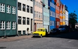 A row of colorful, traditional townhouses lines a street. Each building has multiple windows, and the facades display a variety of colors including green, beige, blue, red, and yellow. Parked along the street is a distinctive yellow vintage car.