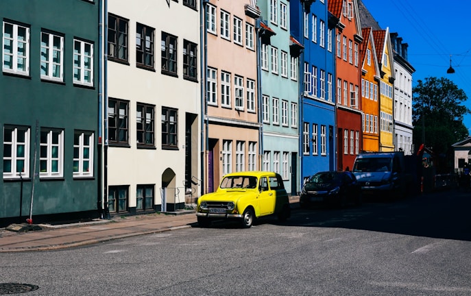 A row of colorful, traditional townhouses lines a street. Each building has multiple windows, and the facades display a variety of colors including green, beige, blue, red, and yellow. Parked along the street is a distinctive yellow vintage car.