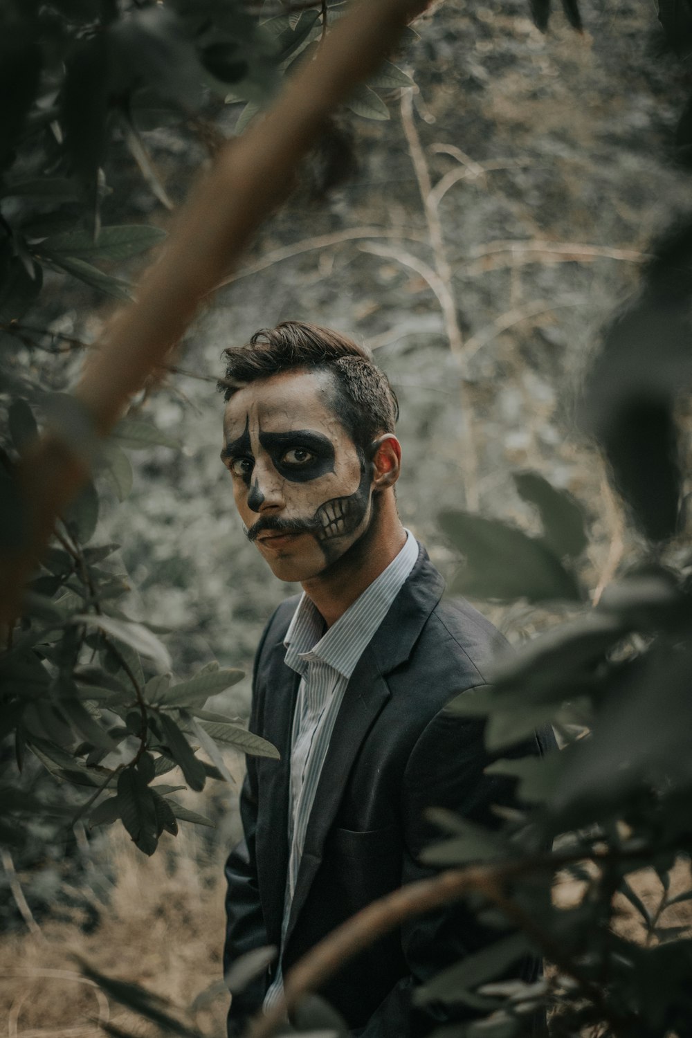 man wearing black notched lapel suit jacket with Halloween face art standing surrounded with tall and green trees