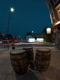 Three wooden barrels are positioned on a sidewalk, chained together. A city street scene unfolds in the background, featuring a bus in motion indicated by blurred light trails and illuminated street lamps casting a soft glow. Buildings are visible, including a sign for 'White Hart Brew Pub' on a building corner.