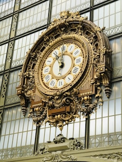 An ornate and large clock with Roman numerals is mounted on a decorative metallic structure against a backdrop of glass windows. The clock features intricate detailing and gold accents.