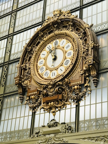 An ornate and large clock with Roman numerals is mounted on a decorative metallic structure against a backdrop of glass windows. The clock features intricate detailing and gold accents.