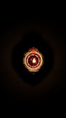 A decorative object resembling a traditional Indian oil lamp is illuminated, centered against a dark background. It features intricate patterns and jewel-like red and green embellishments.