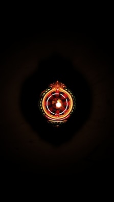 A decorative object resembling a traditional Indian oil lamp is illuminated, centered against a dark background. It features intricate patterns and jewel-like red and green embellishments.