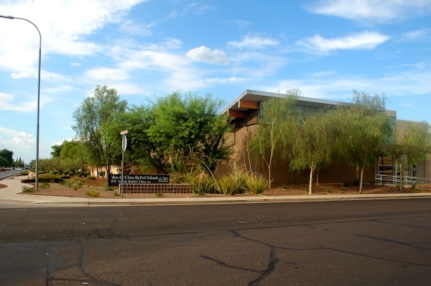 A modern building with a flat roof surrounded by well-maintained landscaping, including trees and shrubs. There's a sign indicating a ballet school, and the scene is set against a clear blue sky with a few clouds. A street and sidewalk are visible in the foreground.
