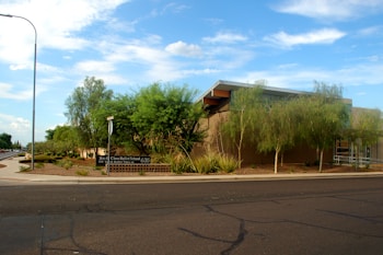 A modern building with a flat roof surrounded by well-maintained landscaping, including trees and shrubs. There's a sign indicating a ballet school, and the scene is set against a clear blue sky with a few clouds. A street and sidewalk are visible in the foreground.