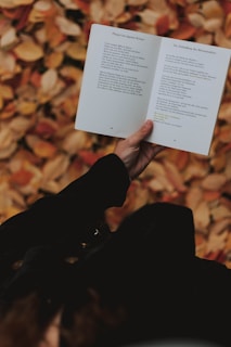 A weathered hand holding a worn copy of the Tao Te Ching against a backdrop of autumn leaves.