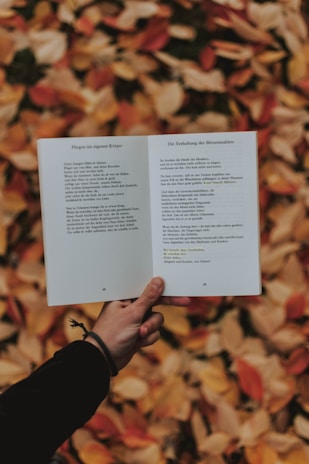 A person holds an open book with German text against a backdrop of autumn leaves in shades of orange, red, and yellow. The person is wearing a black sleeve and a bracelet on the wrist.