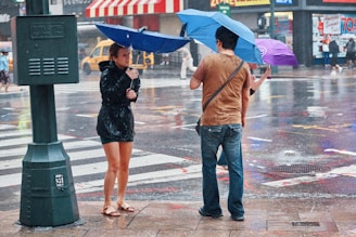 A busy city street scene with people using umbrellas in the rain. The ground is wet with visible reflections of colors and lights. A woman in a black raincoat and sandals stands across from a man in a brown shirt and jeans, both holding blue umbrellas. A third person is partially visible holding a purple umbrella. Yellow taxis can be seen in the background, and a stripy awning adds to the urban setting.