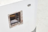 A white wall features a small, square window with a rustic, weathered wooden frame. Inside the window, a portion of a wooden interior and a yellow object are visible. A vintage radio with a long antenna is hanging from the window, and an industrial-style outdoor light fixture is mounted above it.