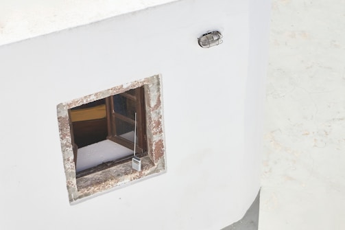 A white wall features a small, square window with a rustic, weathered wooden frame. Inside the window, a portion of a wooden interior and a yellow object are visible. A vintage radio with a long antenna is hanging from the window, and an industrial-style outdoor light fixture is mounted above it.