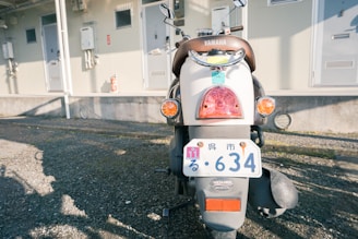 A Yamaha scooter is parked on a gravel surface in front of a building with several utility boxes and a fire extinguisher mounted on the wall. The scooter is seen from the rear, displaying a Japanese license plate and a small inspection sticker. The scene is lit by natural sunlight casting some shadows on the ground.
