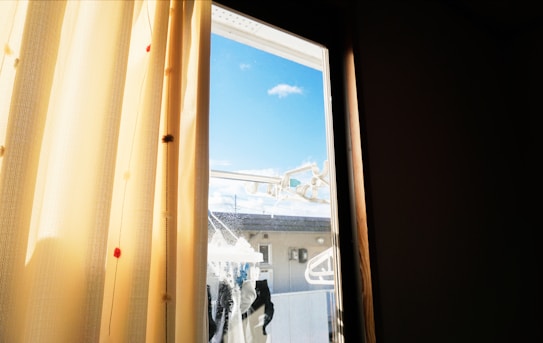 Light yellow curtains partly cover a window with a clear view outside. Sunlight filters through the fabric, creating a warm glow. Outside, there is a blue sky scattered with a few clouds. Several clothes hangers and possibly a laundry rack are visible.