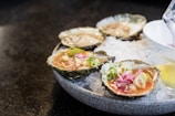 A plate of fresh oysters served on a bed of crushed ice, garnished with green onions and sliced pink pickled ginger. A small container with sauce is placed beside the oysters, and a lemon wedge is partially visible.