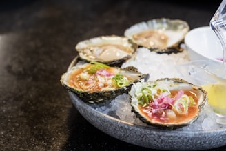 A plate of fresh oysters served on a bed of crushed ice, garnished with green onions and sliced pink pickled ginger. A small container with sauce is placed beside the oysters, and a lemon wedge is partially visible.