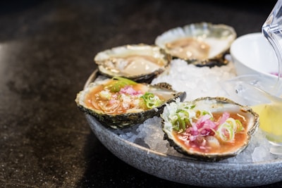 A plate of fresh oysters served on a bed of crushed ice, garnished with green onions and sliced pink pickled ginger. A small container with sauce is placed beside the oysters, and a lemon wedge is partially visible.