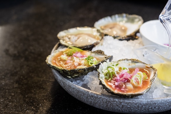 A plate of fresh oysters served on a bed of crushed ice, garnished with green onions and sliced pink pickled ginger. A small container with sauce is placed beside the oysters, and a lemon wedge is partially visible.