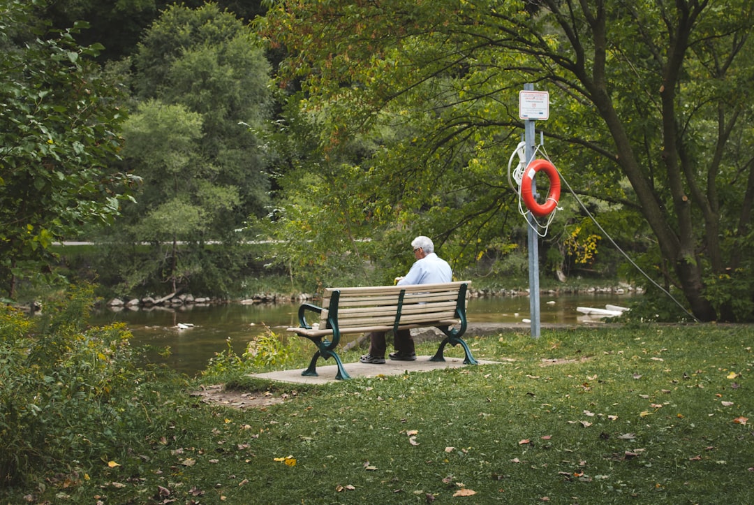 man sitting on wooden bench facing body of water surrounded with tall and green trees during daytime