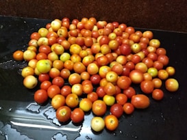 A large pile of small, round, red and yellow tomatoes is spread out on a dark, wet surface. The tomatoes vary slightly in size and color, ranging from vibrant red to light yellow. Droplets of water are visible on some of the tomatoes as well as on the surface, suggesting they have been recently washed.